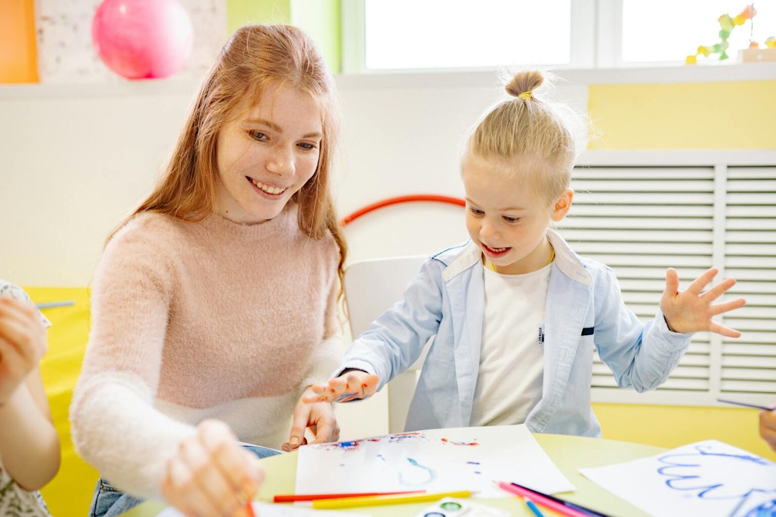 Child engaging in painting activity assisted by smiling teacher in a bright classroom.